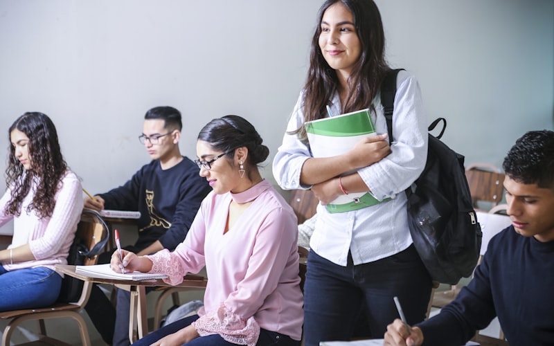 En classe, une élève debout tient un classeur vert dans ses bras, à côté de camarades assis qui prennent des notes